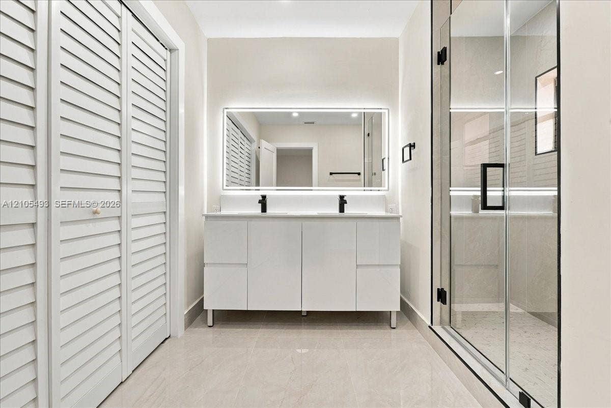 Double vanity with white cabinetry, rectangular vessel sinks, matte black faucets, and a backlit mirror, featuring a walk-in glass-enclosed shower and white louvered closet doors