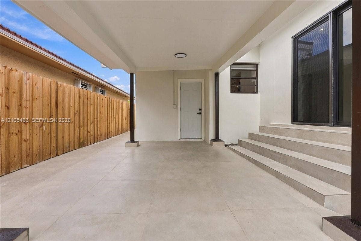 Covered patio featuring large-format porcelain tile flooring, structural support columns, a paneled exterior entry door, and a set of tiered exterior stairs leading to a glass sliding door