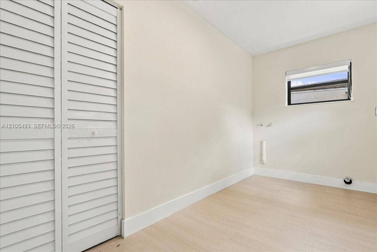 Neutral-toned interior room featuring wood-finish flooring, white louvered closet doors, a horizontal window with integrated roller shade, and wall-mounted utility connections