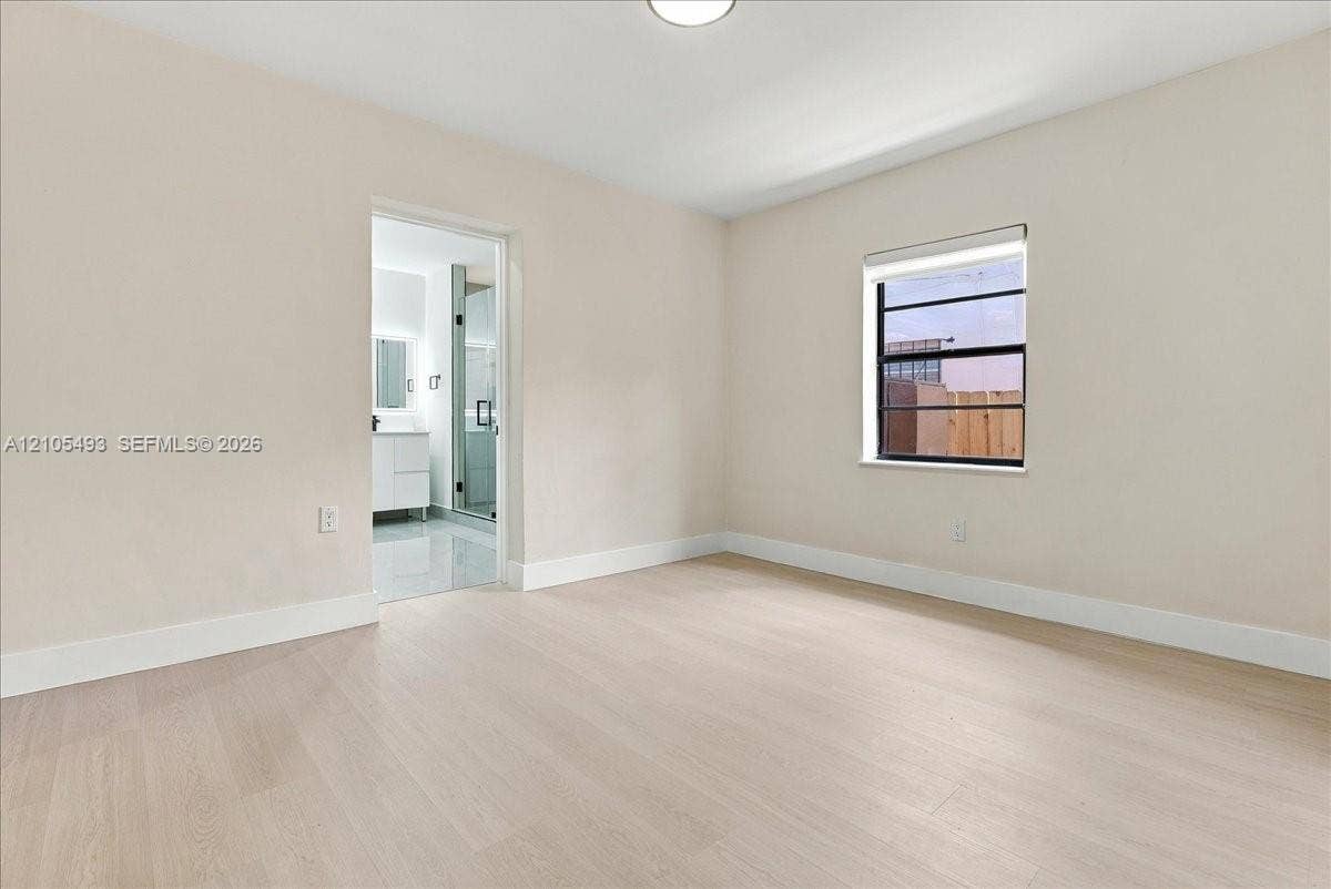 Neutral-toned interior space featuring light-colored wood-finish flooring, a single-pane window, and an entrance to an en-suite with a vanity and glass-enclosed shower