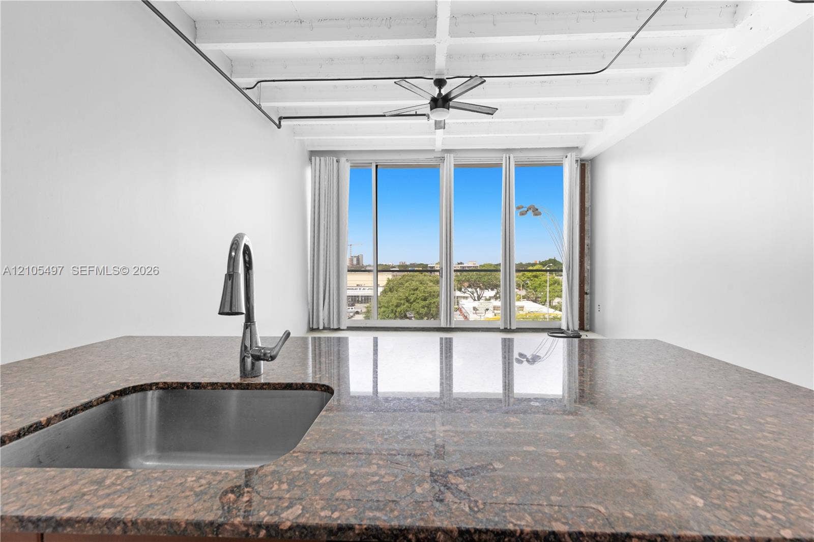 Integrated stainless steel sink with a high-arc faucet, granite-finish countertop, exposed ceiling beams, and full-height windows