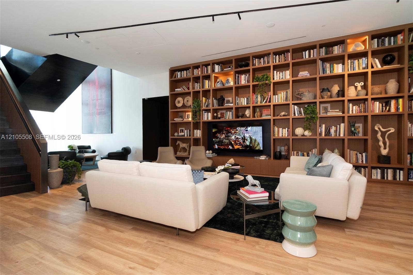 Expansive living area featuring wood-finish flooring, a full-height built-in bookshelf wall with integrated television, and a dark-finished open staircase