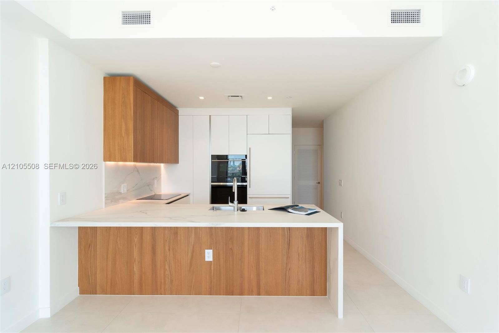 Kitchen featuring contemporary wood-finish and white cabinetry, built-in stainless steel appliances, a large island with an undermount sink, and light-toned flooring