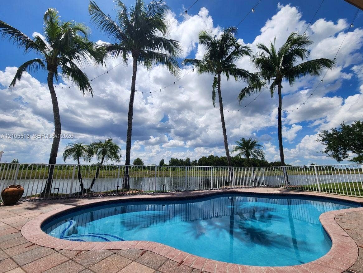 Curved-edge swimming pool with mosaic tile details, surrounded by paver decking, white picket fencing, and mature palm trees