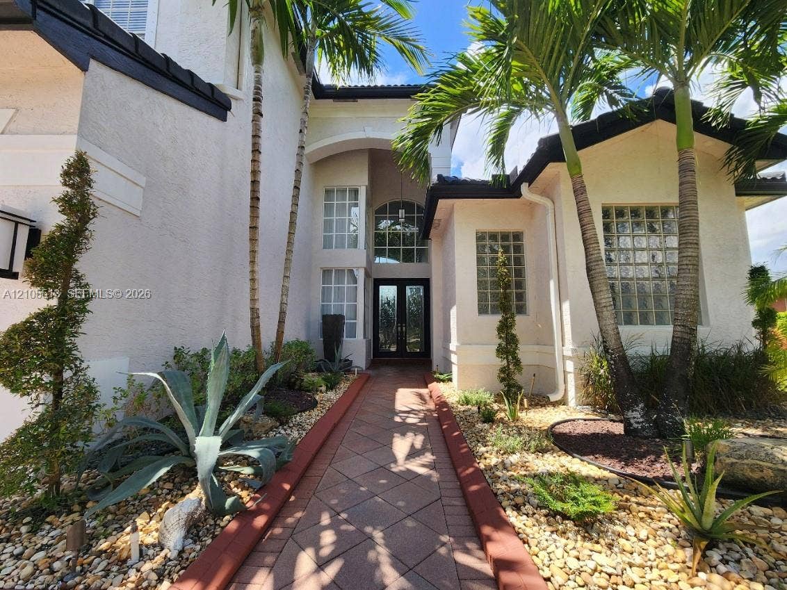 Stucco exterior with a tiled roof, arched entry, double-height windows, and a paved walkway, complemented by mature palm trees and landscaped rock gardens