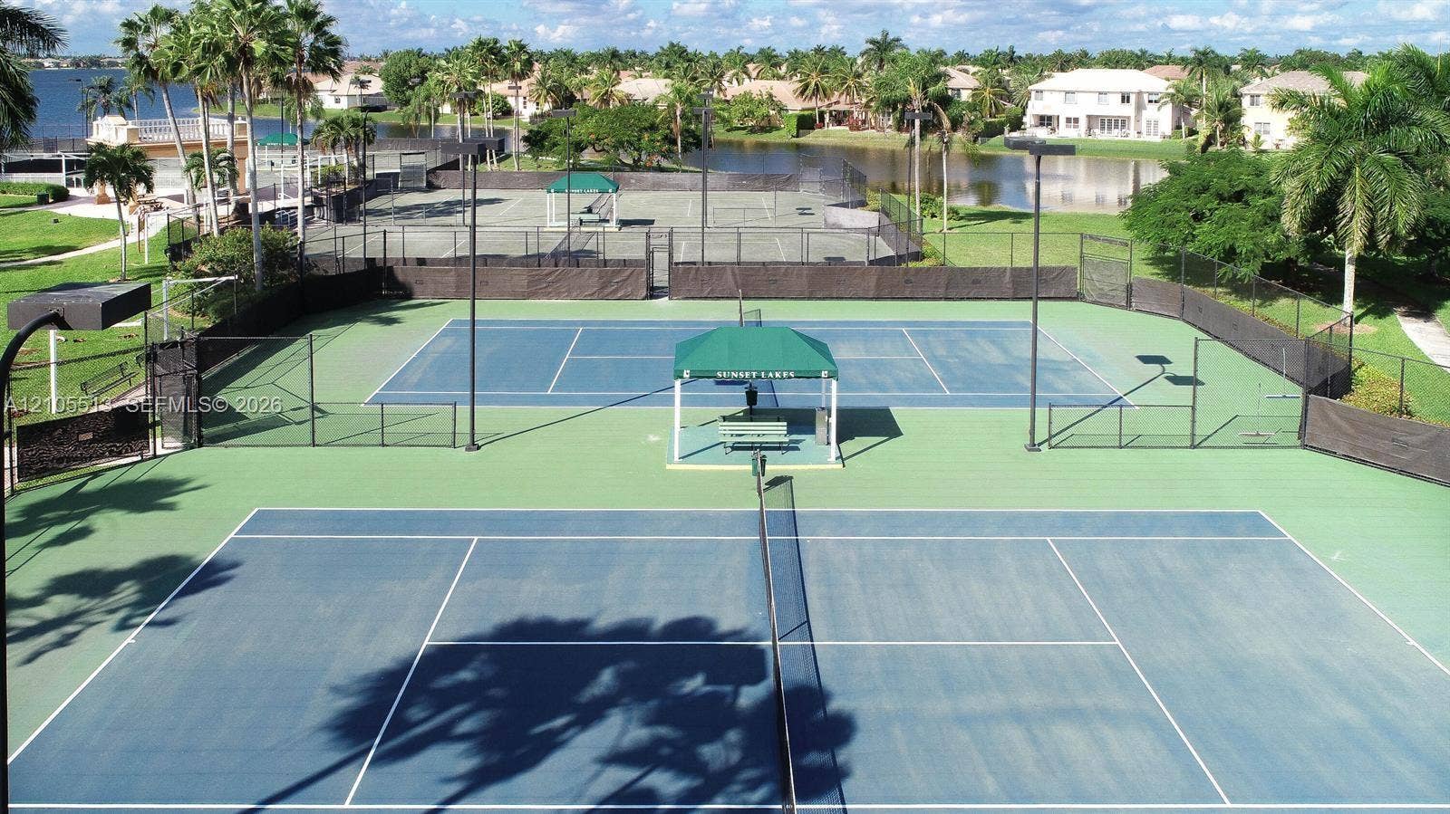 Multi-court tennis facility featuring blue and green acrylic surfaces, perimeter chain-link fencing, high-mast lighting, and a central shaded observation gazebo