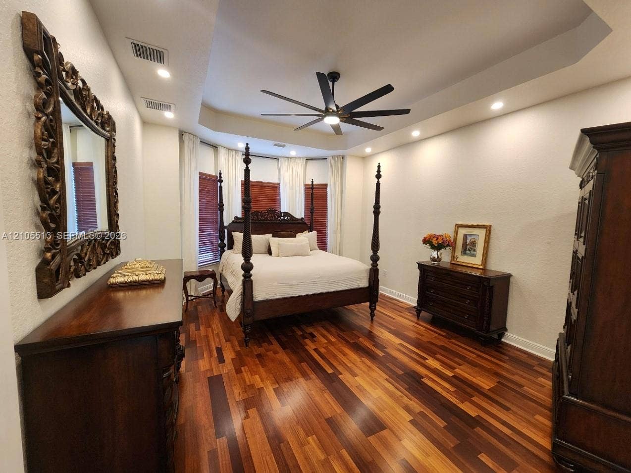 Expansive primary bedroom with hardwood flooring throughout, tray ceiling, recessed lighting, ceiling fan, and large windows with wood privacy blinds