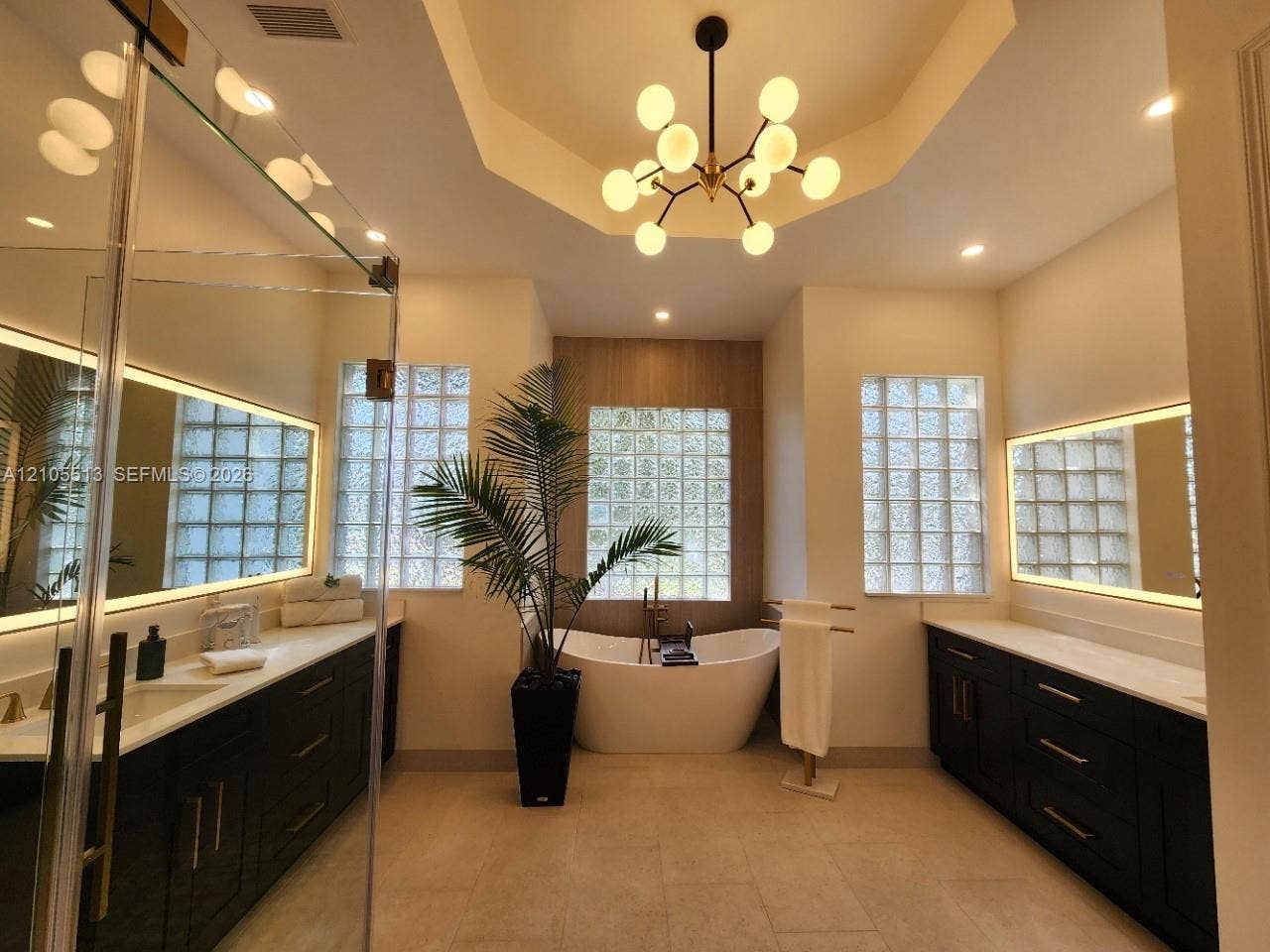 Spacious newly remodeled spa-inspired primary bathroom featuring porcelain tile, a freestanding soaking tub, dual custom vanities with quartz counters, recessed lighting, glass block windows, lighted mirrors and a tray ceiling with a modern chandelier