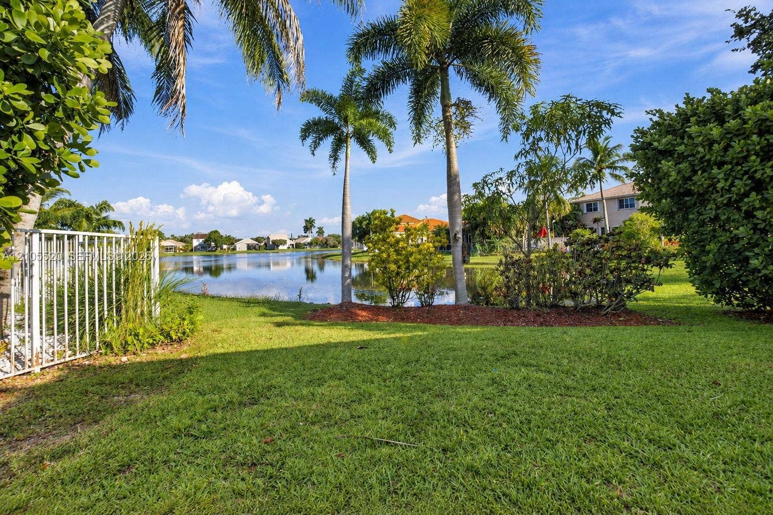 649 Willow Bend Road, Weston, Weston, FL 33327 Waterfront backyard featuring a manicured lawn, established palm trees, decorative mulch beds, and white metal perimeter fencing