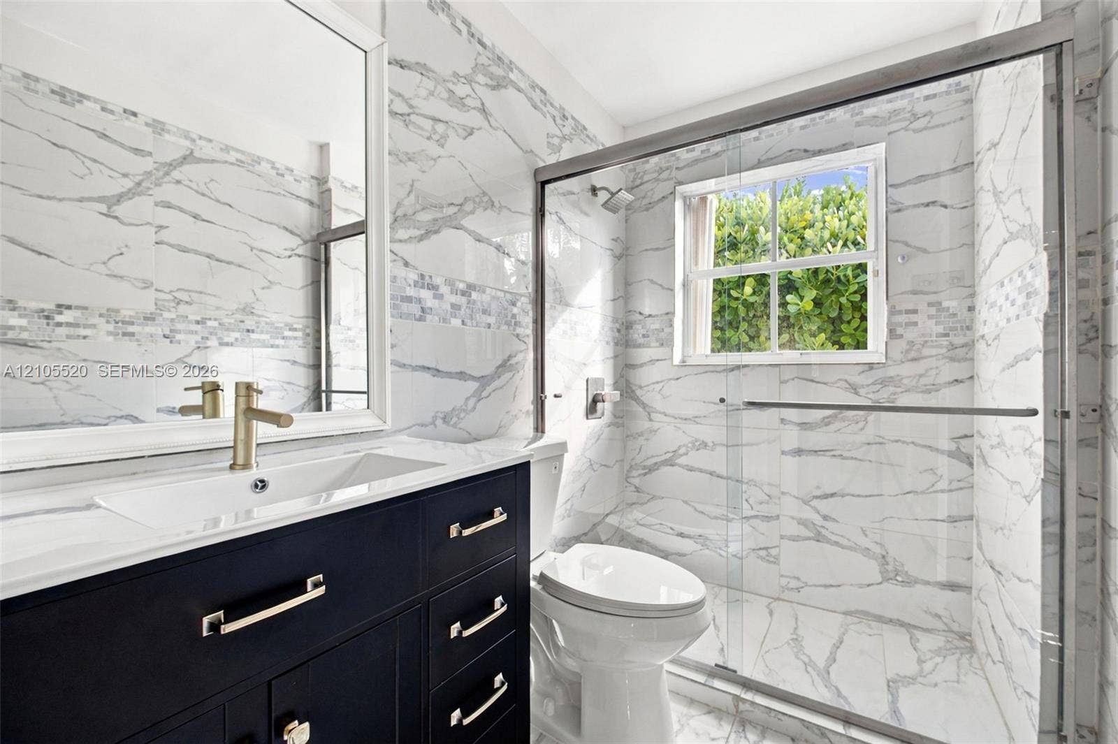 649 Willow Bend Road, Weston, Weston, FL 33327 Dark navy vanity cabinet with metallic hardware, white countertop, large-format veined tile wall surfacing, glass-enclosed shower, and framed vanity mirror