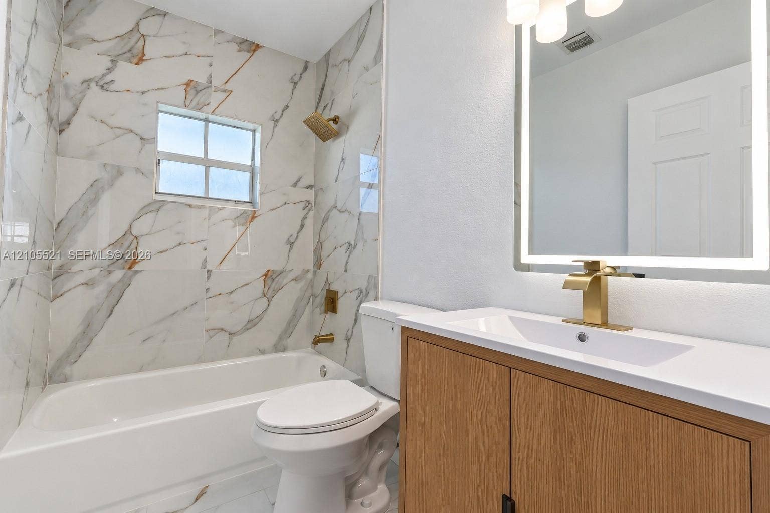 Bathroom featuring large format marble-look wall tile, a built-in bathtub, a vanity with wood-finish cabinetry, a white countertop, and a lighted mirror