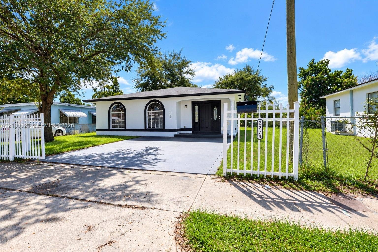 White exterior with arched windows, dark trim, a black front door, a paved driveway, and a white picket fence