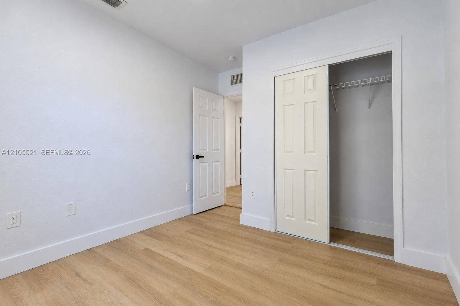 Light wood-finish flooring with white baseboards, a white paneled door with a black handle, and a white sliding closet door
