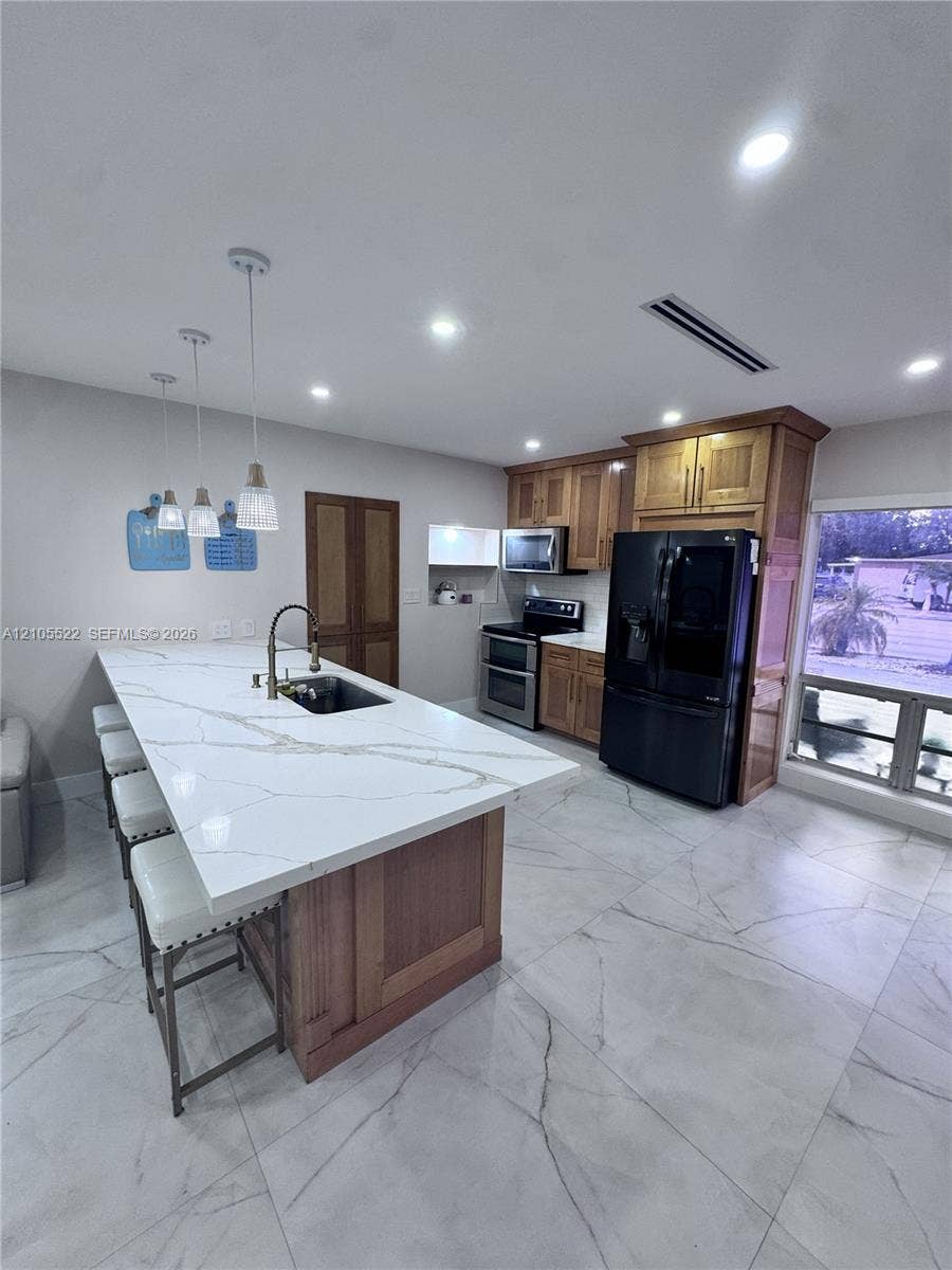 Kitchen featuring an expansive island with a stone-finish countertop, wood-finish cabinetry, stainless steel sink with gooseneck faucet, and integrated black appliances