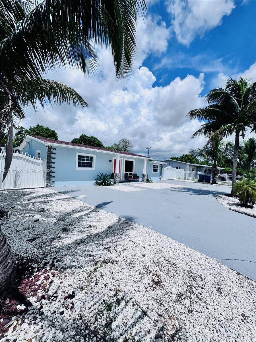 Light blue exterior with white trim, a red entry door, a white privacy fence, an asphalt driveway, and white gravel landscaping