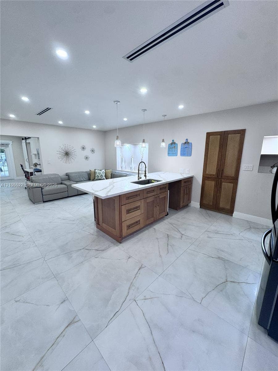 Spacious open-concept room featuring large format tile flooring, a kitchen island with a white countertop and integrated sink, pendant lighting, recessed ceiling lights, and wooden double doors