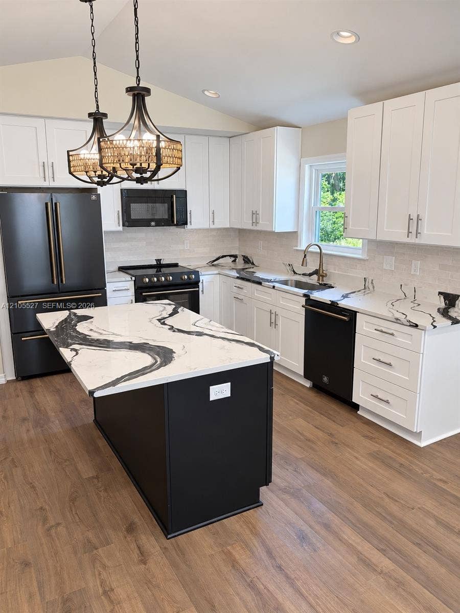 Kitchen featuring white shaker cabinetry, black appliances, a central island with a waterfall countertop, wood-finish flooring, and two decorative pendant lights