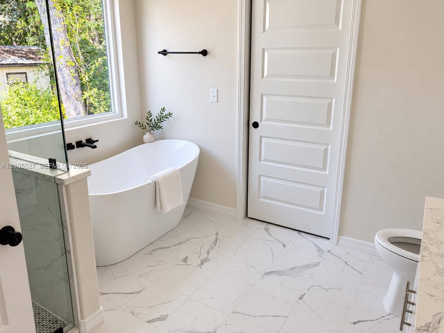 Freestanding soaking tub with matte black fixtures, framed window, and marble-look floor tiling
