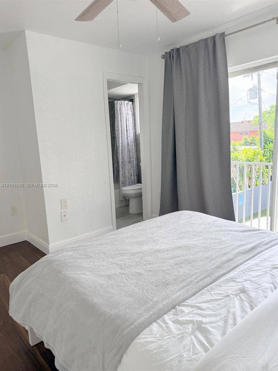 Wood-finish flooring, ceiling fan, sliding glass door, white balcony railing, and a bathroom entrance with a toilet and shower curtain