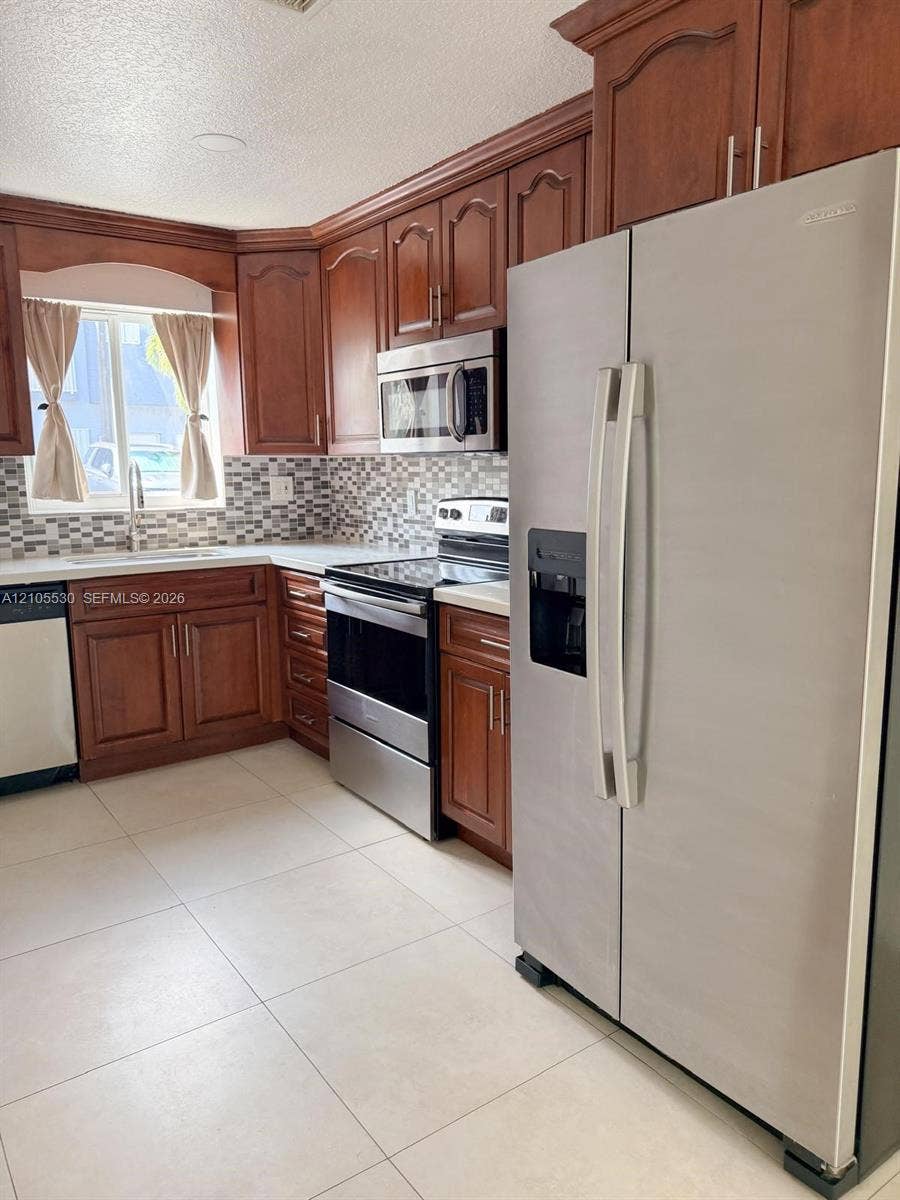 Kitchen featuring wood cabinetry, stainless steel appliances, mosaic tile backsplash, white countertops, and tile flooring