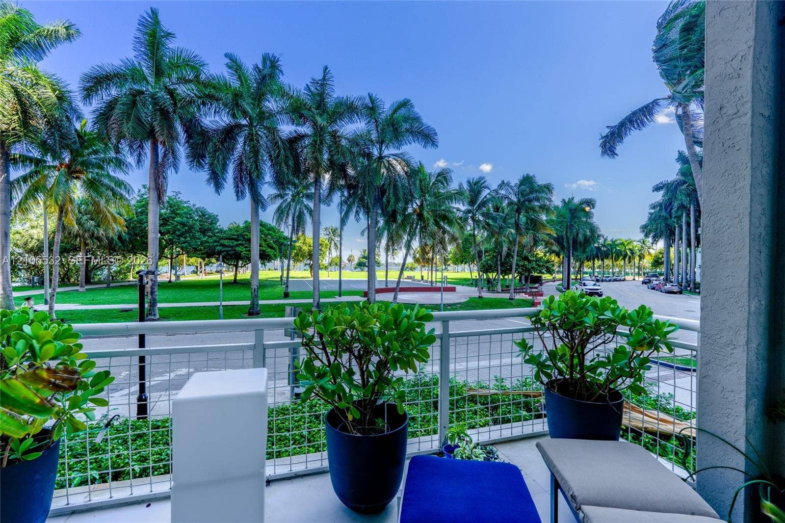 Covered balcony featuring white metal railings and concrete flooring, overlooking a green space with mature palm trees