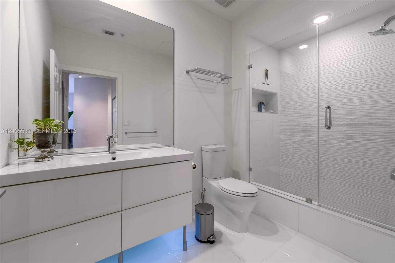 Bathroom featuring a gloss-finish vanity with integrated sink, large wall-mounted mirror, walk-in shower with glass enclosure and textured tile surround, and recessed lighting