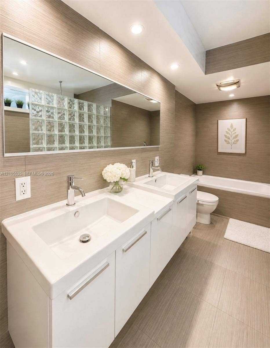 Dual vanity with integrated sinks and chrome fixtures, large rectangular mirror, recessed lighting, and a glass block shower wall