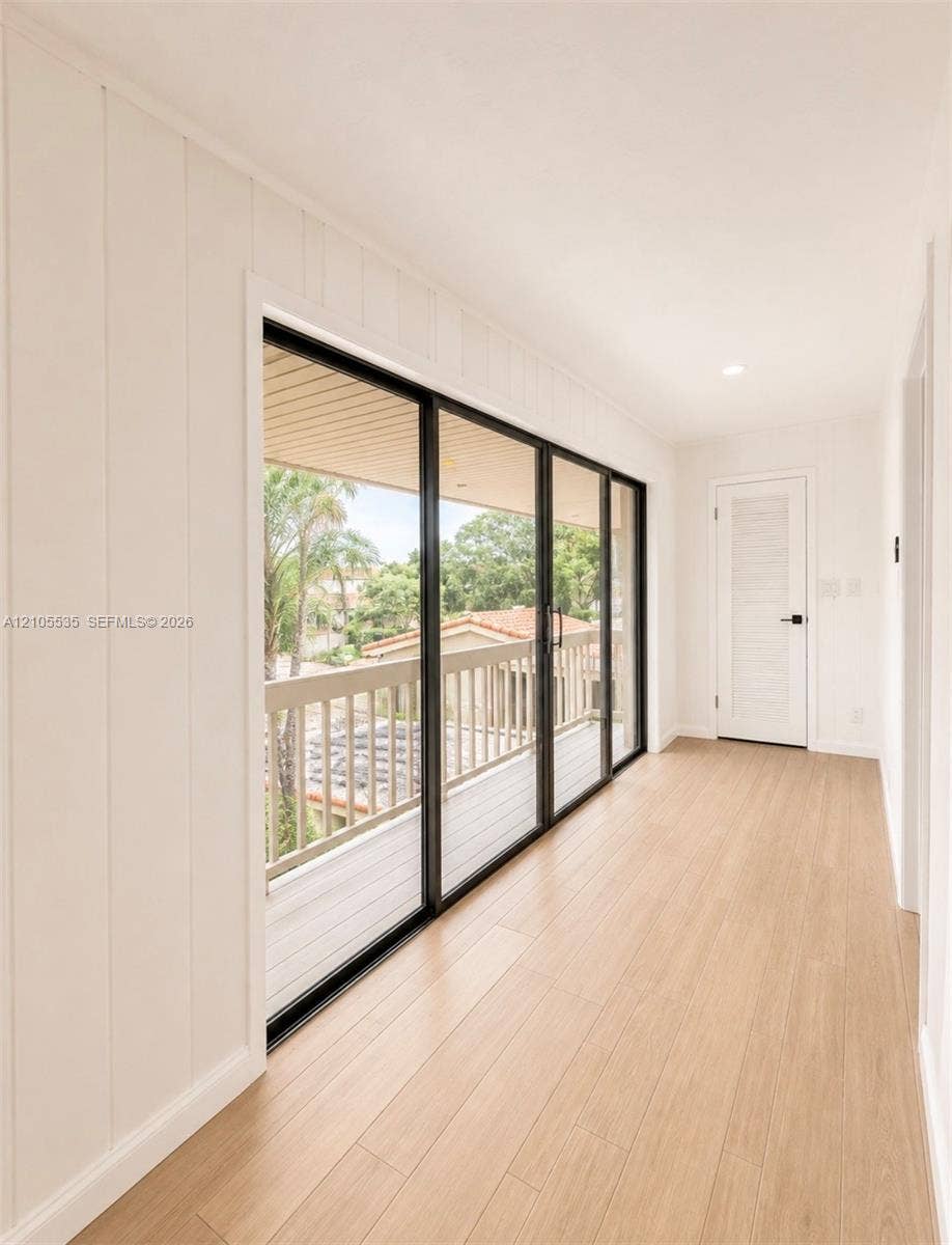 Recessed lighting, wood-finish flooring, white paneled walls, white paneled door with black hardware, and multi-panel sliding glass doors accessing an outdoor balcony with a wooden railing