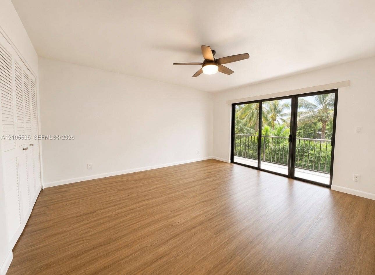 Wood-finish flooring throughout, louvered bi-fold closet doors, ceiling fan with integrated lighting, and sliding glass doors to a balcony with metal railing