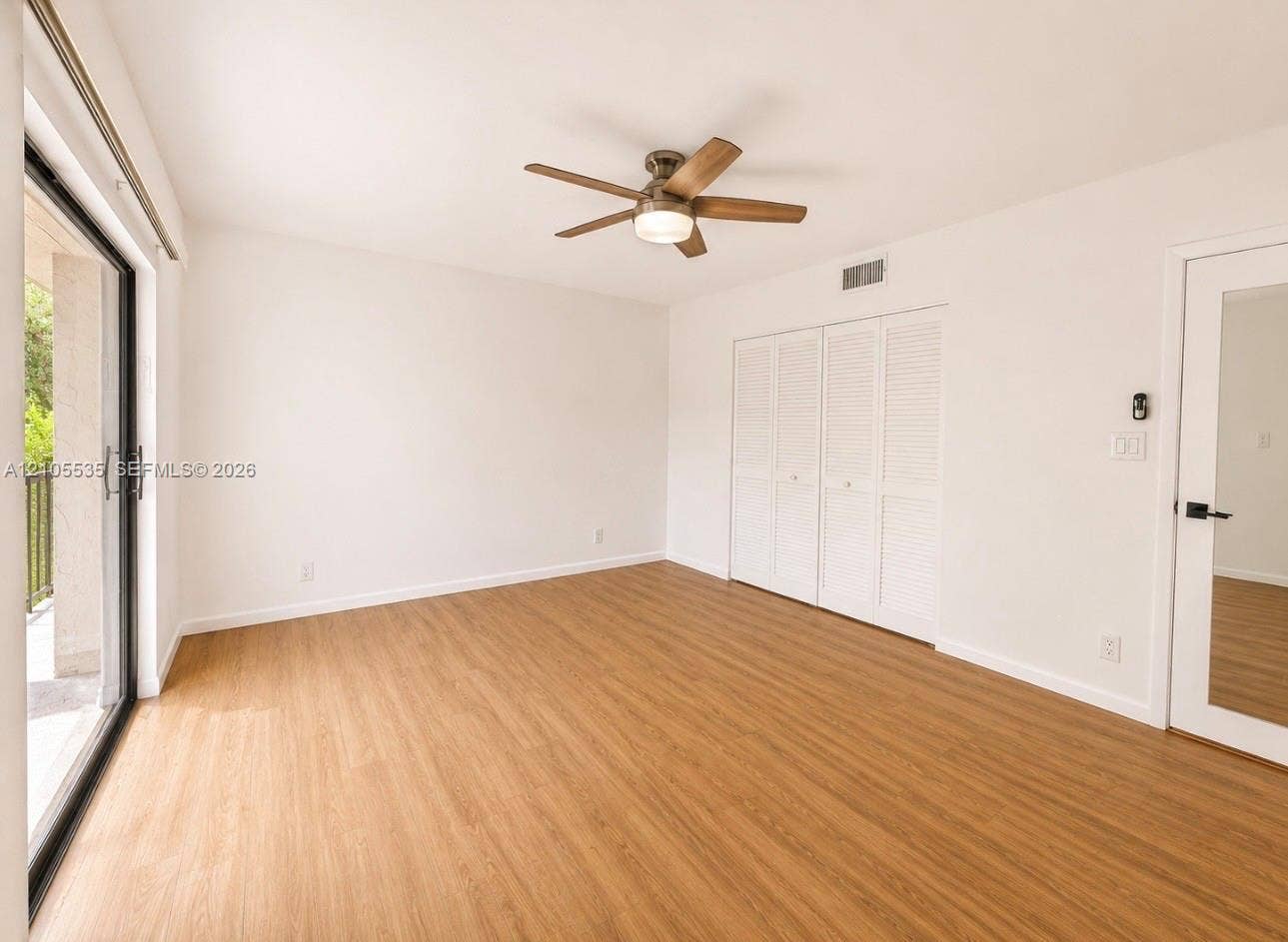 Wood-finish flooring throughout, featuring a ceiling fan with integrated lighting, sliding glass door with balcony, louvered bi-fold closet doors, and a paneled door with frosted glass