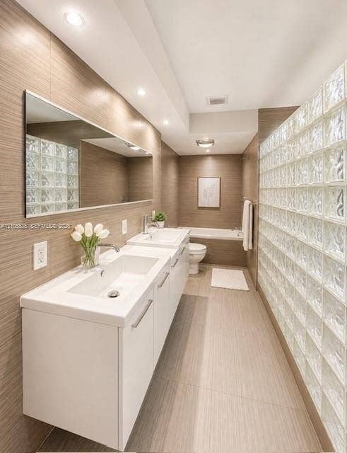 Dual floating vanities with integrated sinks, full-width mirror, recessed lighting, glass block wall, and tiled flooring