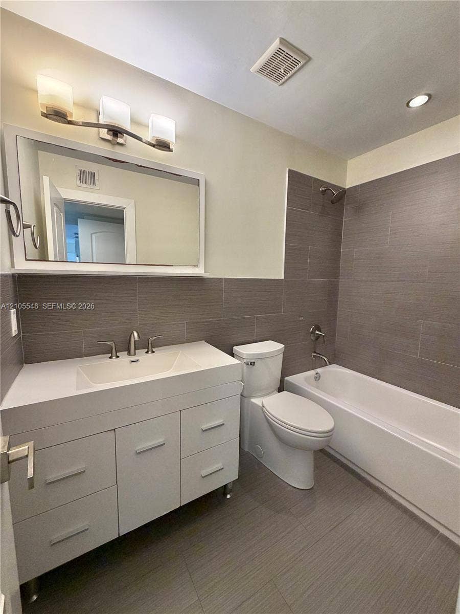 White vanity with rectangular integrated sink, three-light wall-mounted fixture, large framed mirror, and floor-to-ceiling gray tiled shower surround