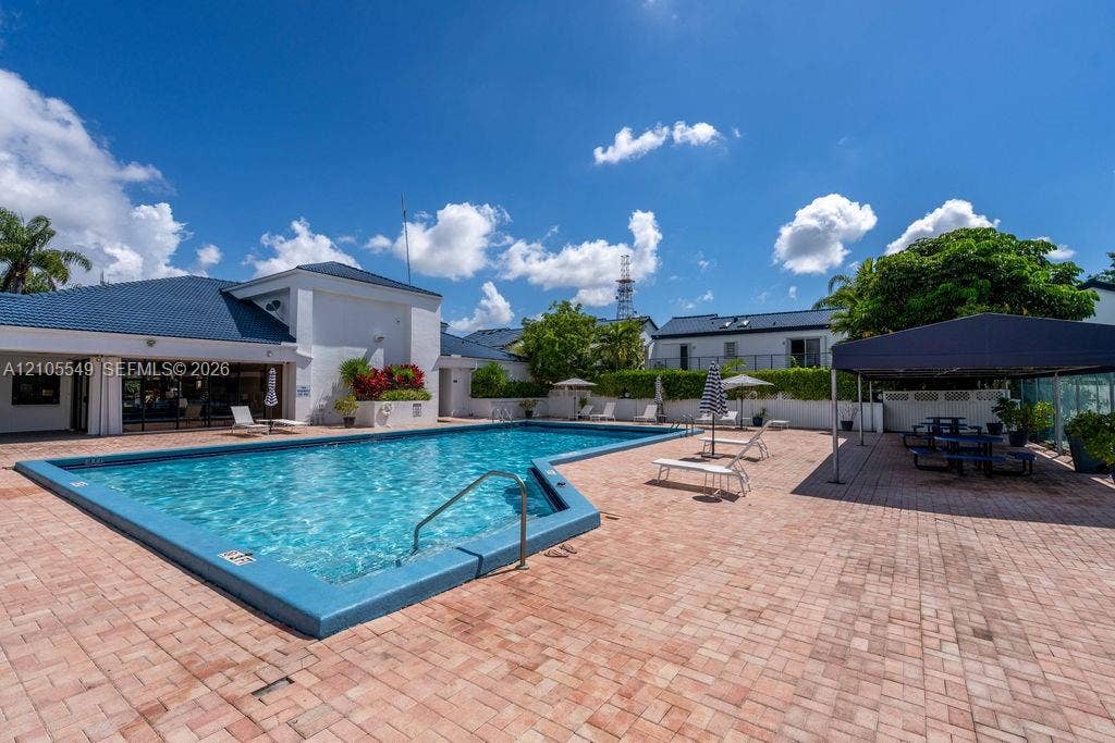 Expansive outdoor pool with a blue tile edge, surrounded by a large brick paver deck, permanent poolside canopy, and lush perimeter landscaping