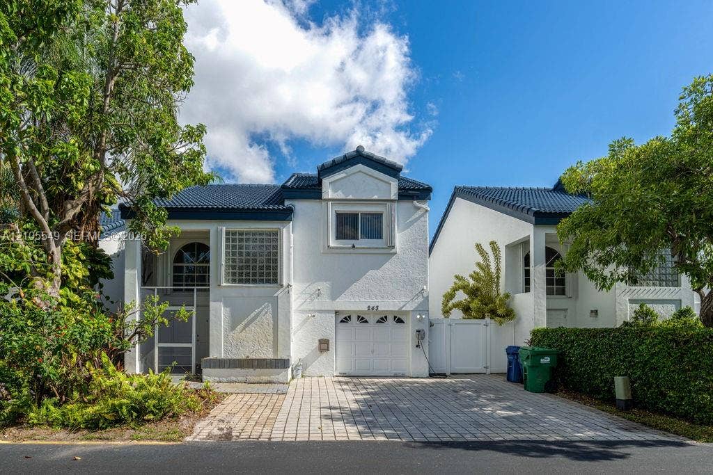 White exterior with a dark-tiled roof, featuring a paver driveway, an attached garage, and a screened entry