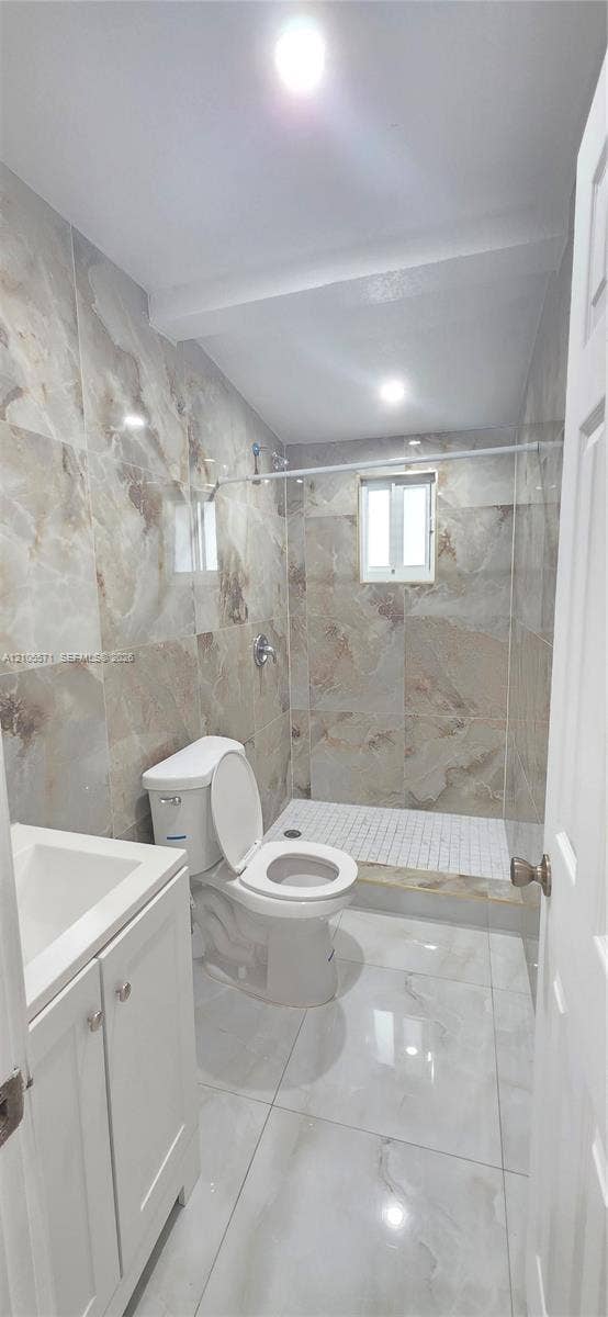 Bathroom featuring full-height stone-look tile, a walk-in shower with a glass panel, a white vanity with an integrated sink, and recessed lighting