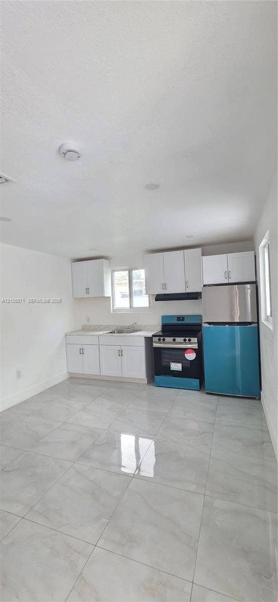 White cabinetry with stainless steel hardware, a stainless steel range with an overhead ventilation hood, a stainless steel and teal refrigerator, and a stainless steel sink with a window