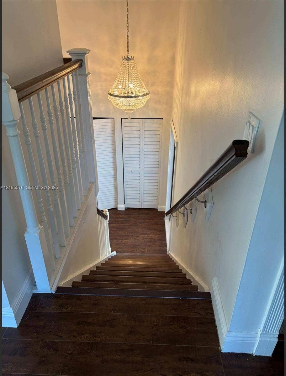 Staircase featuring dark wood-finish flooring, white spindle railings, a wood-finish handrail, crystal chandelier, and louvered double-door closets