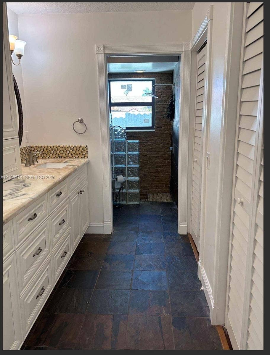 Bathroom featuring a white cabinetry vanity, stone-patterned countertop with a mosaic tile backsplash, slate-finish floor tile, and a textured stone wall in the shower area