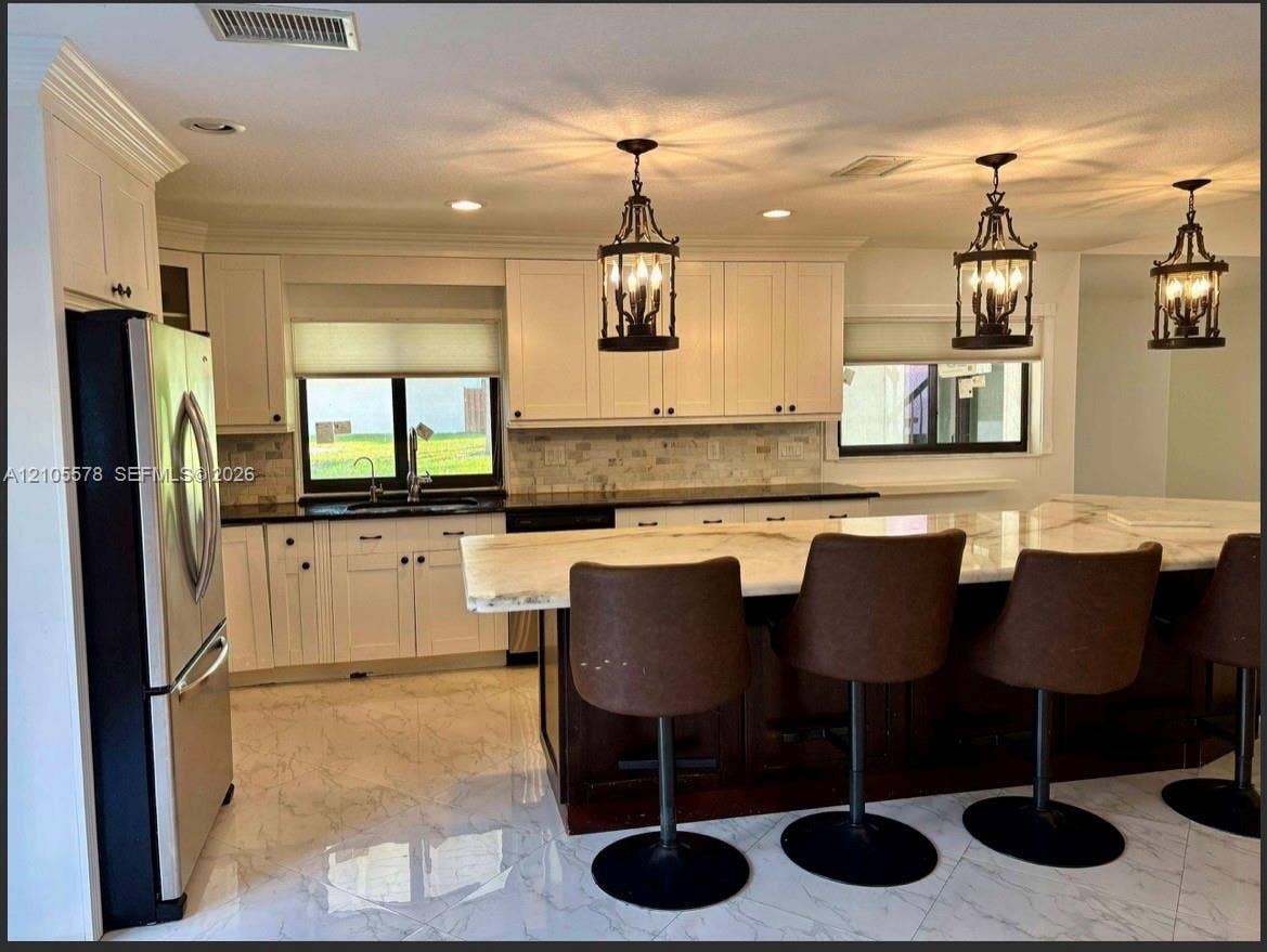 Kitchen featuring white shaker cabinetry, stone-topped central island with seating, stone backsplash, stainless steel appliances, and polished tile flooring