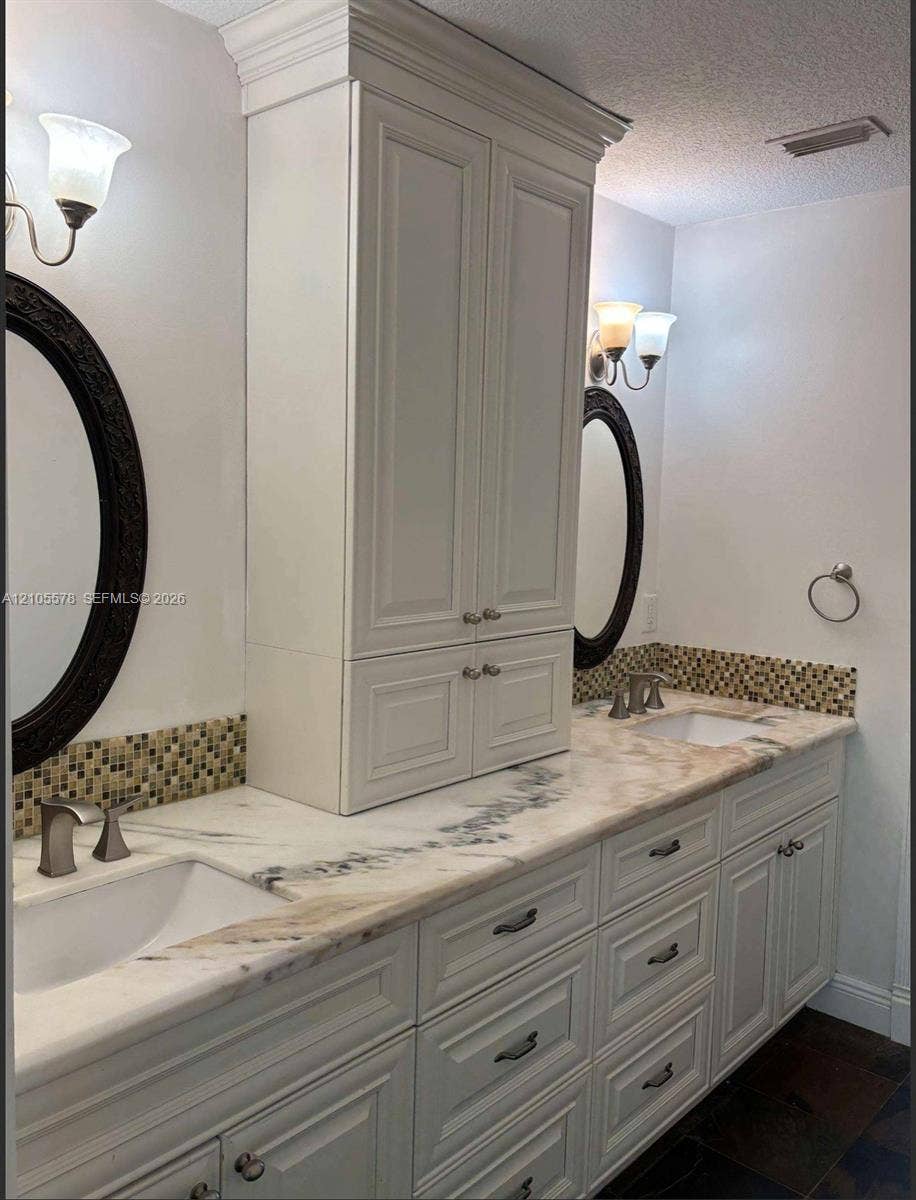 Double vanity with a stone countertop, white shaker-style cabinetry with crown molding, dual undermount sinks, and decorative mosaic tile backsplash