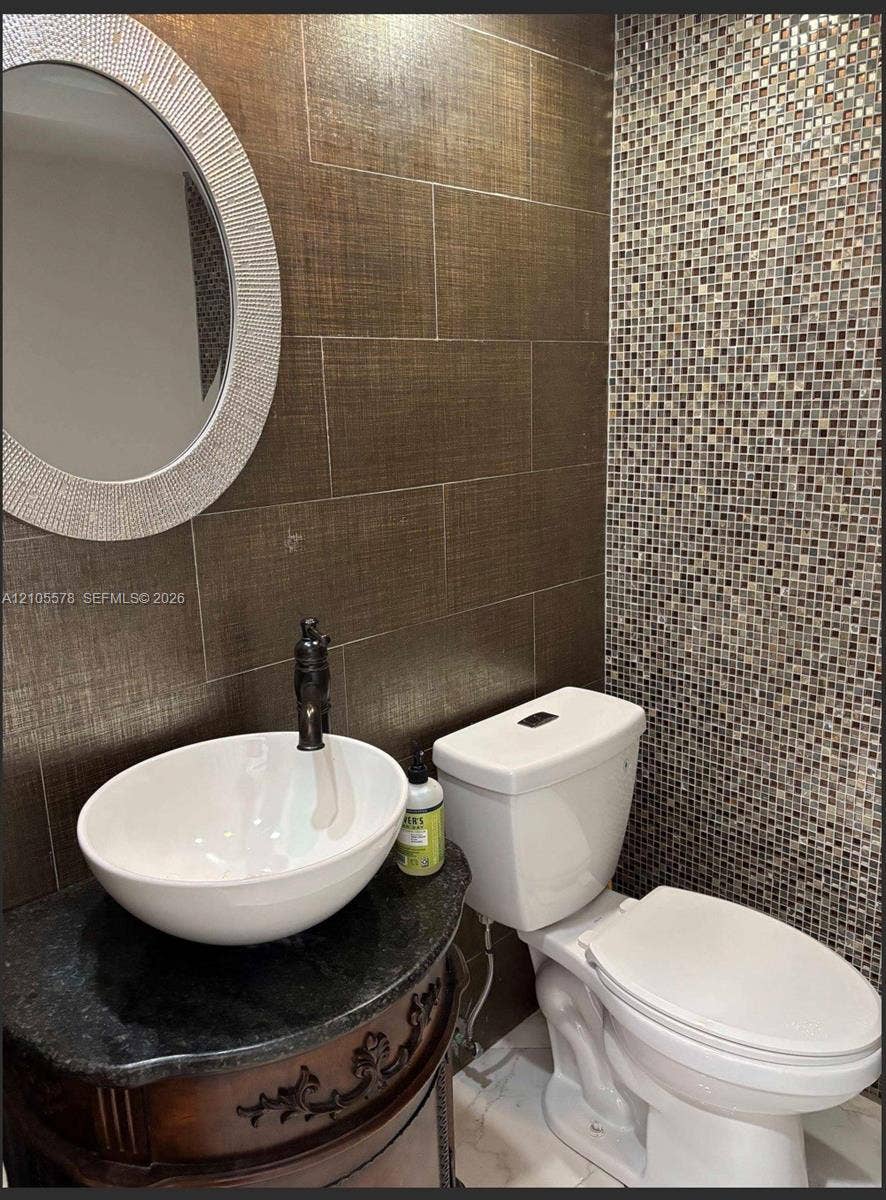 Oval vessel sink with a dark-finish faucet, stone-top vanity, textured wall tiling, and decorative mosaic accent wall