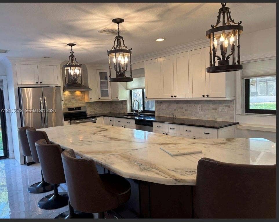 Kitchen featuring a large central island with light-toned natural stone surfacing, white cabinetry, dark countertops, tiled backsplash, and decorative lantern-style pendant lighting
