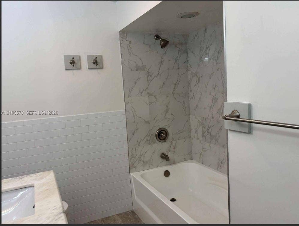 Bathtub with stone-finish tile surround, white subway tile wainscoting, integrated countertop vanity, and metal fixtures