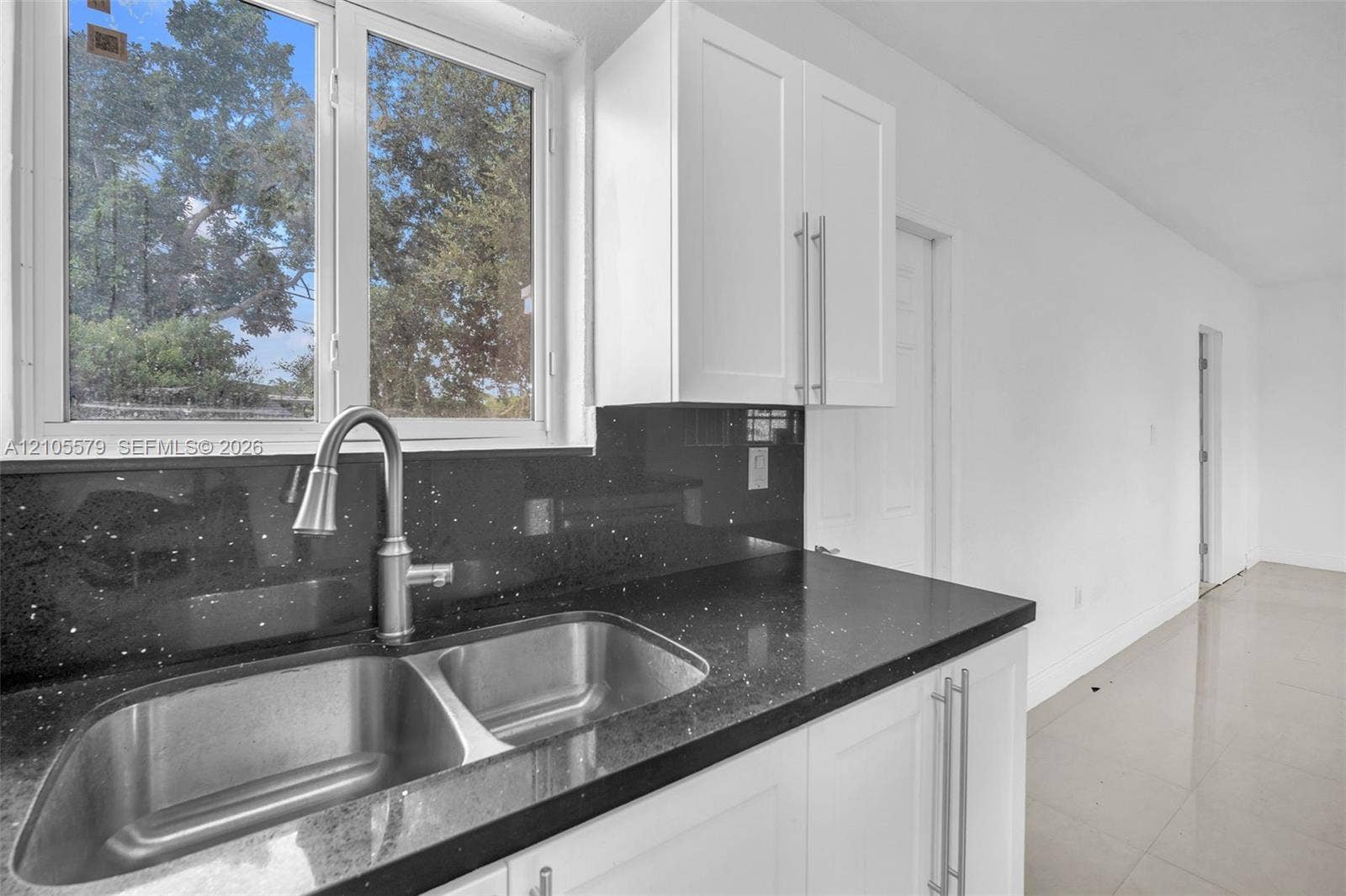 1750 NW 185th Street, Miami Gardens, FL 33056 Kitchen area featuring white shaker-style cabinetry, dark stone-finish countertops, stainless steel dual-basin sink, high-arc brushed nickel faucet, and large-format tile flooring