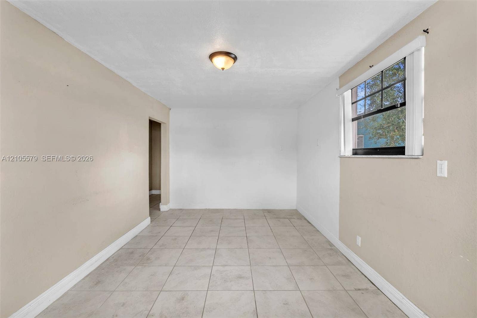 1750 NW 185th Street, Miami Gardens, FL 33056 Neutral-toned interior space featuring light-colored tile flooring, smooth walls, a single-pane window with dark muntins, and a flush-mount ceiling light fixture