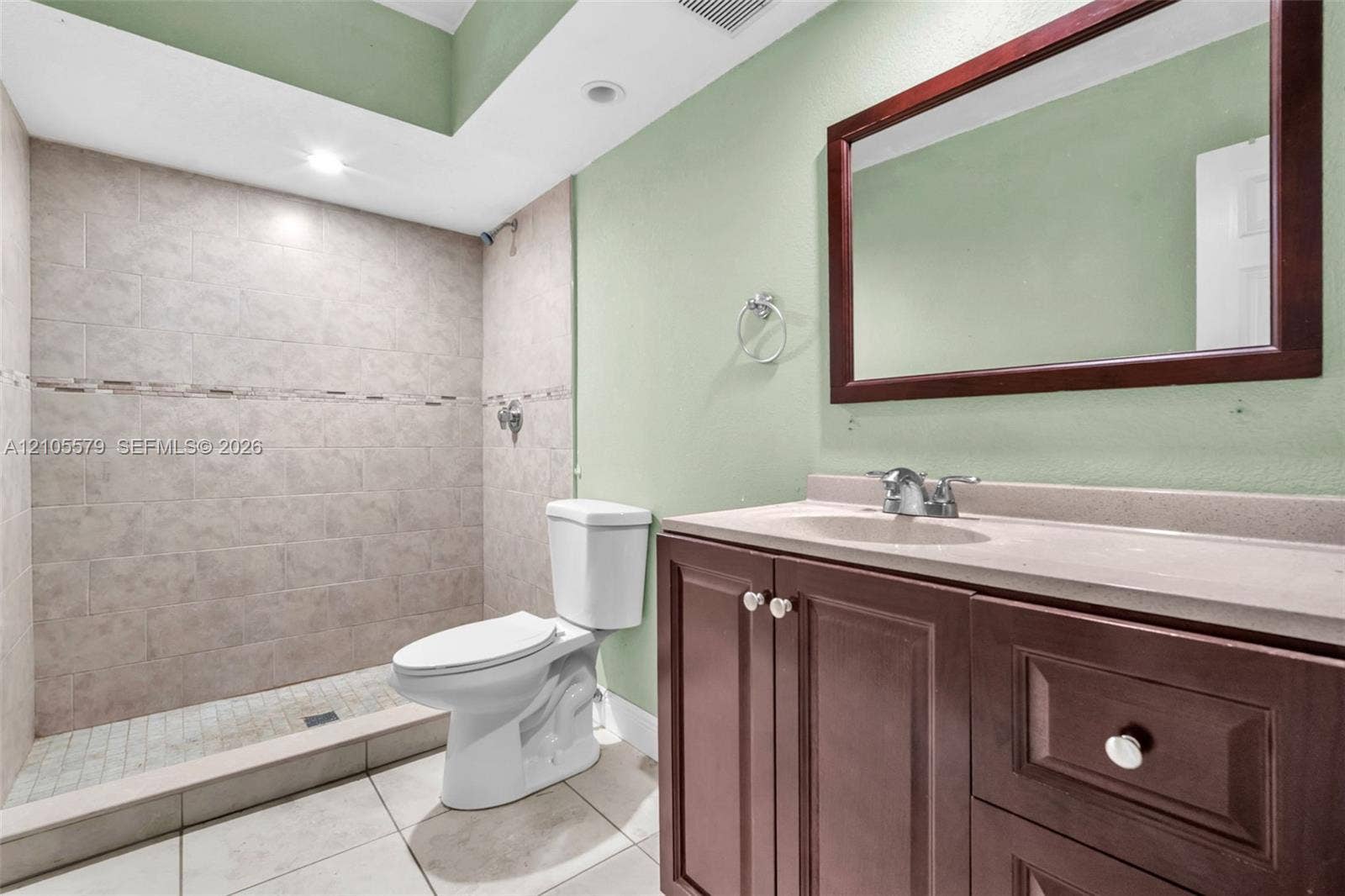 1750 NW 185th Street, Miami Gardens, FL 33056 Bathroom featuring a walk-in tiled shower with an accent border, vanity with integrated stone-finish countertop, dark wood-finish cabinetry, and a framed wall-mounted mirror