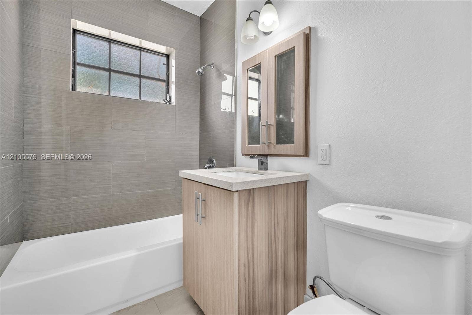 1750 NW 185th Street, Miami Gardens, FL 33056 Bathroom featuring a tile-surround bathtub with a rectangular window, light wood-finish vanity cabinet, stone-composite countertop, and a double-door medicine cabinet