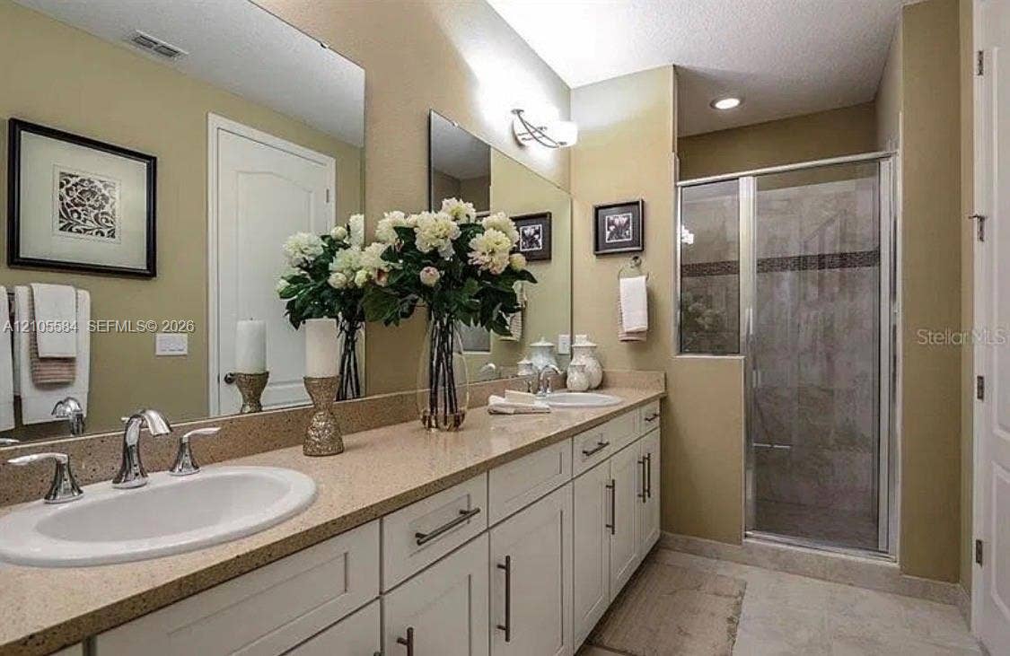 10197 Eagle Creek Center Boulevard, Orlando, FL 32832 Dual-sink vanity with light-tone countertops, white cabinetry with silver-tone hardware, large wall-mounted mirrors, and a glass-enclosed shower with tiled surround