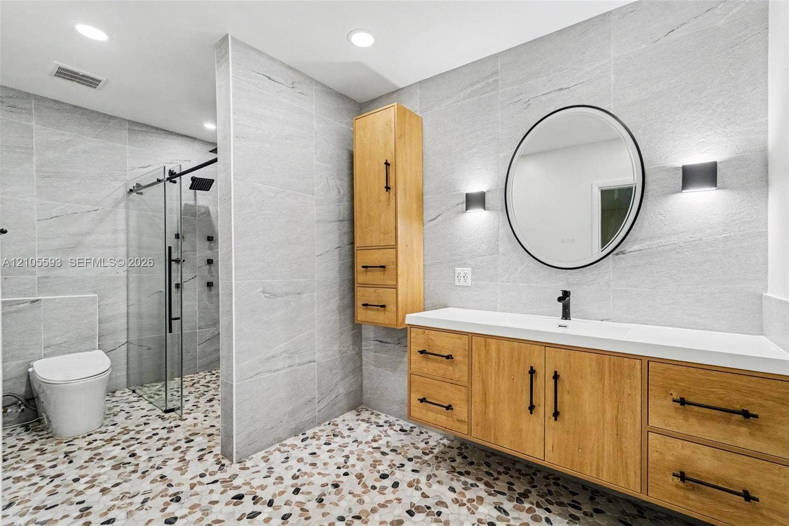 597 Grace Street, Sebastian, FL 32958 Wall-mounted wood-finish vanity with a rectangular sink, walk-in glass shower enclosure, stone-tiled walls, integrated cabinetry, and round vanity mirror