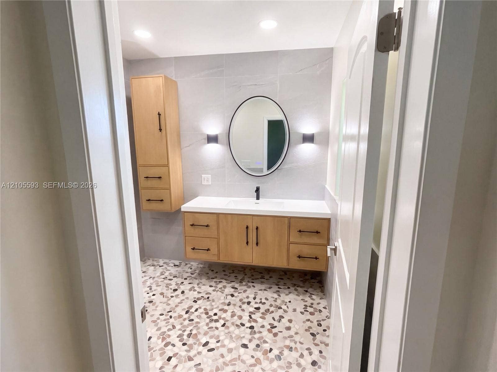 597 Grace Street, Sebastian, FL 32958 Floating wood-finish vanity with matte black hardware, integrated rectangular sink, wall-mounted oval mirror, light-toned stone-patterned wall tile, and pebble-textured floor tile
